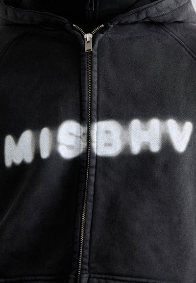 Schwarze Kapuzenjacke mit einem strukturierten Stoff, featuring aufgesprühten weißen Schriftzug "MISBHV" über der Brust, silbernes Reißverschlussdetail.