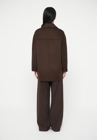 Cappotto oversize di misto lana marrone con colletto a revers, silhouette dritta e tre bottoni su ogni polsino. Abbinato a pantaloni marroni a gamba larga.