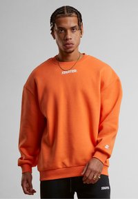 Oransje oversized hettegenser laget av bomull, med en hvit "STARTER"-logo på brystet og ribbede mansjetter og avslutning.