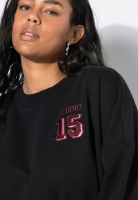 Zwarte sweatshirt met geborduurd "STUDIO 15" in bordeauxrood en metallic zilver. Zachte textuur met een ronde halslijn en een losse fit.