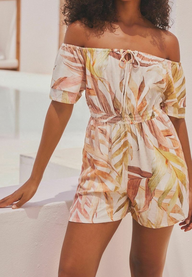 Combinaison à épaules dénudées ornée de fleurs, à manches courtes, fabriquée en tissu léger. Elle possède une taille avec cordon de serrage et un motif de feuilles tropicales dans des couleurs douces.