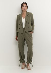 Alyvuogių žalia pritaikyta švarkas su dvigubais mygtukais, derinamas su atitinkamomis krovinių kelnėmis, turinčiomis raištelių apykakles, ir balta V formos iškirptės palaidine.