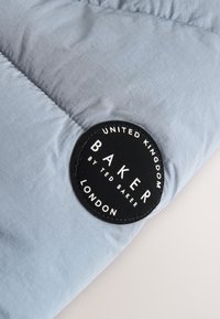 Tela acolchada azul claro con una parche circular negra que dice "United Kingdom Baker by Ted Baker London."