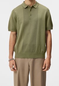Polo à manches courtes vert olive avec un motif à rayures verticales texturées, un col classique et un ourlet côtelé. Porté avec un pantalon beige.