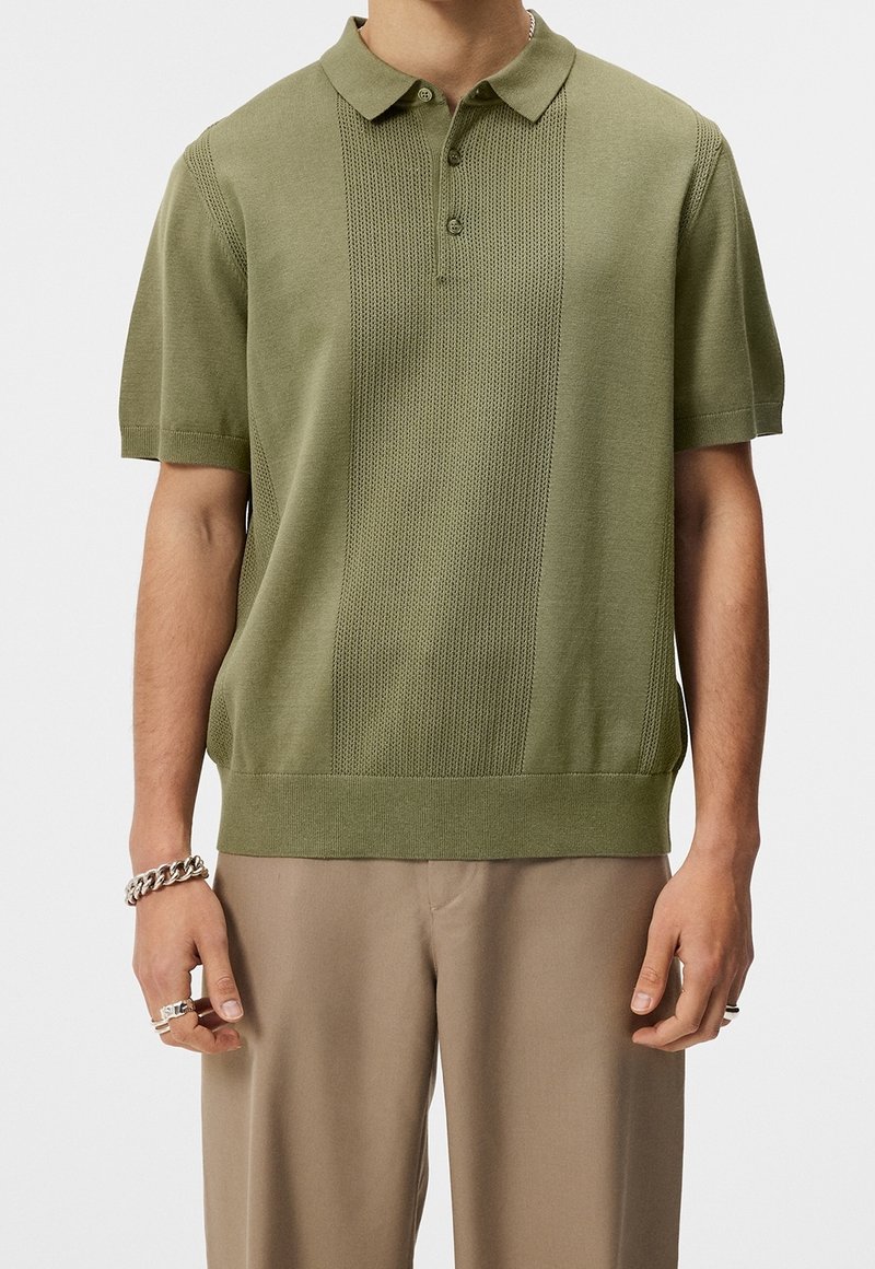Polo à manches courtes vert olive avec un motif à rayures verticales texturées, un col classique et un ourlet côtelé. Porté avec un pantalon beige.