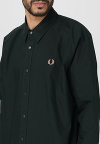 Camicia button-up verde scuro realizzata in tessuto testurizzato, con colletto a punta e un piccolo logo ricamato in rosa sul petto.