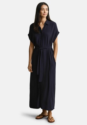 Femme portant une robe-chemise bleu marine ceinturée à manches courtes, debout avec une main dans la poche et des sandales marron sur fond blanc.