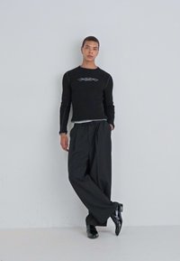 Cămașă neagră cu mânecă lungă, decorată cu un design grafic argintiu, asociată cu pantaloni negri largi și pantofi negri strălucitori. Siluetă simplă și modernă.