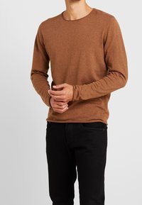 T-shirt marron à manches longues avec un col rond et des poignets ajustés, porté avec un pantalon noir. Le tissu semble doux et texturé.