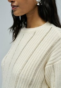 Crèmekleurige geribde trui met metallic zilveren strepen, ronde halslijn en een structuur breiwerk. Heeft een relaxte pasvorm en zichtbare stikseldetails.