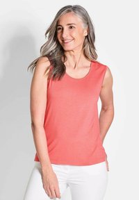 Korallfarbener ärmelloser Top mit Rundhalsausschnitt, aus weichem Stoff, mit glatter Textur und schlichtem Design. Über weißen Hosen getragen.