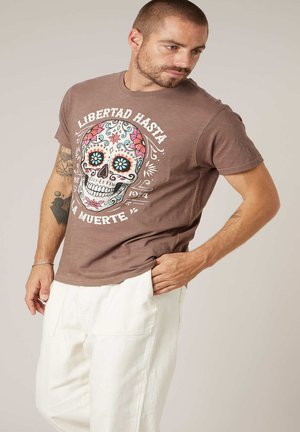 Man met tatoeages die een bruin t-shirt draagt met een kleurrijke suikerschedel en Spaanse tekst, gecombineerd met witte broek, staand tegen een effen achtergrond.