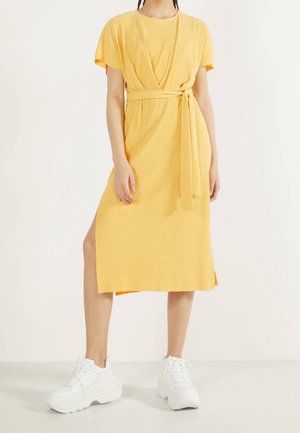 Vrouw die een gele midi wikkeljurk met korte mouwen draagt en witte chunky sneakers, staand tegen een effen lichte achtergrond.