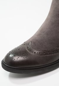 Gros plan sur la pointe d'une chaussure brogue en cuir marron foncé et daim, avec des motifs décoratifs perforés et une semelle noire.