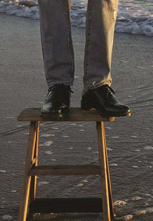 Zwarte leren schoenen op een houten kruk, geplaatst op nat zand met watergolven. Spijkerbroek met vervaagde gedeelten en lichte slijtage.