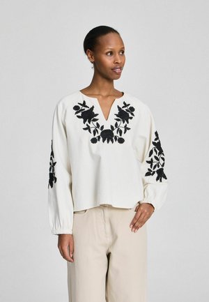 Kobieta ubrana w białą bluzkę z długim rękawem z czarną kwiatową haftowaną na piersiach i rękawach, w połączeniu z beżowymi spodniami, patrząca w bok.