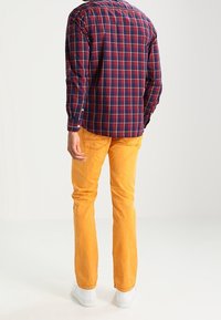 Rotes und marineblau kariertes Langarmhemd, kombiniert mit orangen, gerade geschnittenen Hosen und weißen Turnschuhen, von hinten gezeigt.