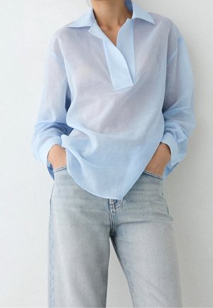 Personne portant une chemise légère bleu clair à manches longues, rentrée lâchement dans un jean en denim clair, les mains dans les poches.