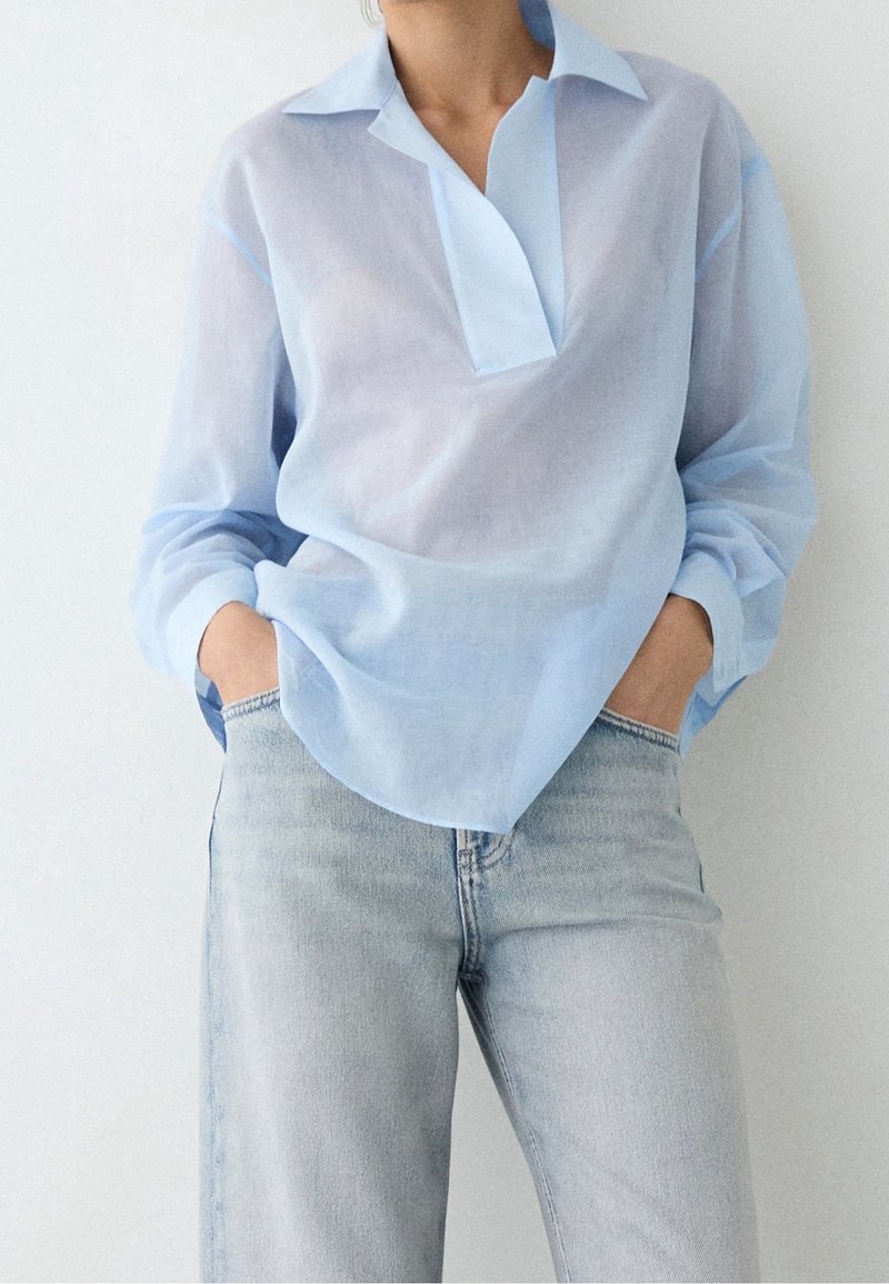 Personne portant une chemise légère bleu clair à manches longues, rentrée lâchement dans un jean en denim clair, les mains dans les poches.