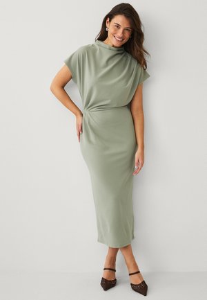 Lachende vrouw die met één hand op haar heup staat, gekleed in een lichtgroene midijurk met korte mouwen en bruine pumps met een spits toelopende neus.