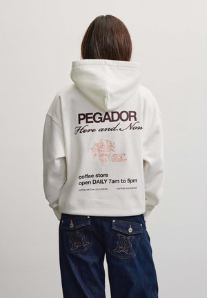 Person iført en hvid hættetrøje med tekst og illustration af en kaffebutik på ryggen, kombineret med mørkeblå jeans med broderede lommer.