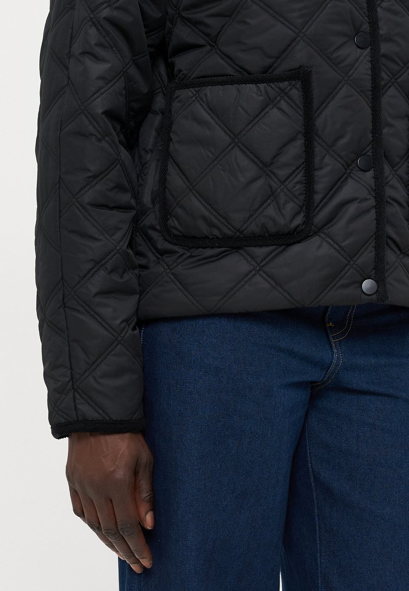 Veste matelassée noire avec un motif en losange, dotée d'une poche à rabat, de bordures sombres et de fermetures à pression, portée par-dessus un jean en denim bleu.