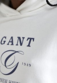 Bílá mikina s kapucí se modrým textem "GANT" a "1949" v klasickém písmu, doplněná o dekorativní písmeno "G". Hladká textura.