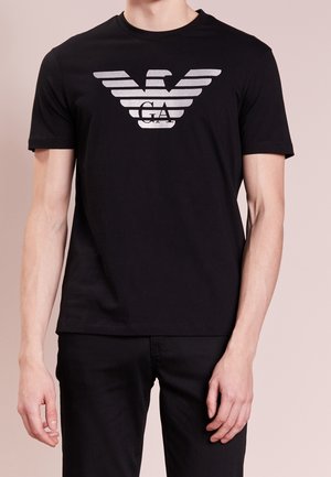 Man draagt een zwart T-shirt met een groot zilverachtig adelaarlogo en de letters GA op de borst, gecombineerd met een zwarte broek.