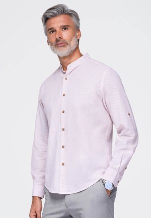 Homme aux cheveux gris et à la barbe portant une chemise à rayures rose clair à boutons et un pantalon gris, debout avec une main dans la poche devant un fond uni.