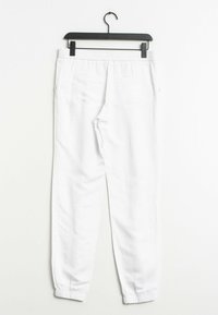 Valkoiset puuvillasekoitetut collegehousut, joissa on joustava vyötärö, sivutaskut ja kuminauhalla varustetut nilkat, roikkumassa mustassa koukussa valkoista taustaa vasten.