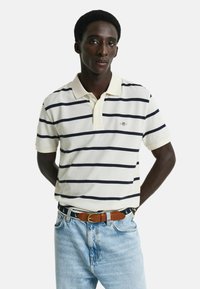 Gestreepte polo in crèmekleur met marineblauwe horizontale strepen, gemaakt van katoen, met korte mouwen en een klassieke kraag. Gecombineerd met lichtblauwe jeans en een bruine riem.