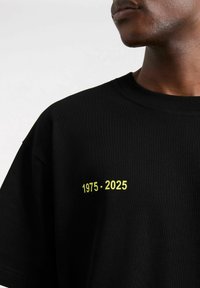 Czarna bawełniana koszulka z krótkim rękawem i okrągłym dekoltem. Napis "1975 - 2025" w jaskrawym żółtym kolorze z przodu po lewej stronie.