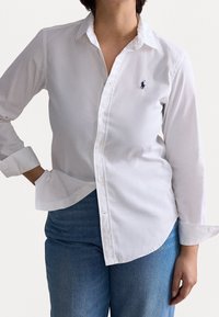 Camisa de cuello blanco hecha de algodón, con un frente de botones y un pequeño logo azul en el lado izquierdo del pecho. Mangas largas con puños remangados.
