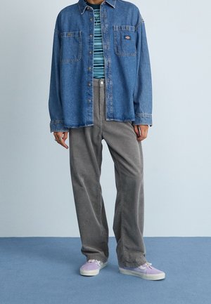 Person iført en oversized denimjakke over en stribet skjorte, grå kordfløjlsbukser og lavendelfarvede sneakers, stående på blå tæppe.