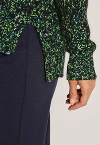 Hand liegt neben einer dunkelblauen Hose, trägt einen schwarz-grün melierten Strickpullover mit geripptem Bündchen und seitlichem Schlitz.
