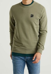 Sudadera verde oliva con cuello y puños acanalados, con un pequeño parche de logo negro en el pecho, combinada con jeans azules.