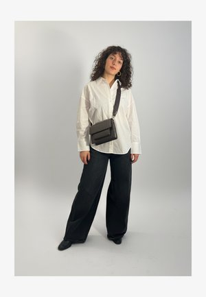 Femme aux cheveux bouclés portant une chemise blanche oversize, un pantalon large noir, des chaussures noires, des boucles d'oreilles créoles dorées et un sac bandoulière noir.