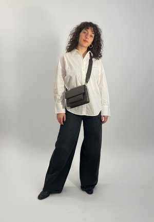 Femeie cu păr creț purtând o cămașă albă supradimensionată, pantaloni negri largi, pantofi negri, cercei mari de aur și o geantă de umăr neagră.