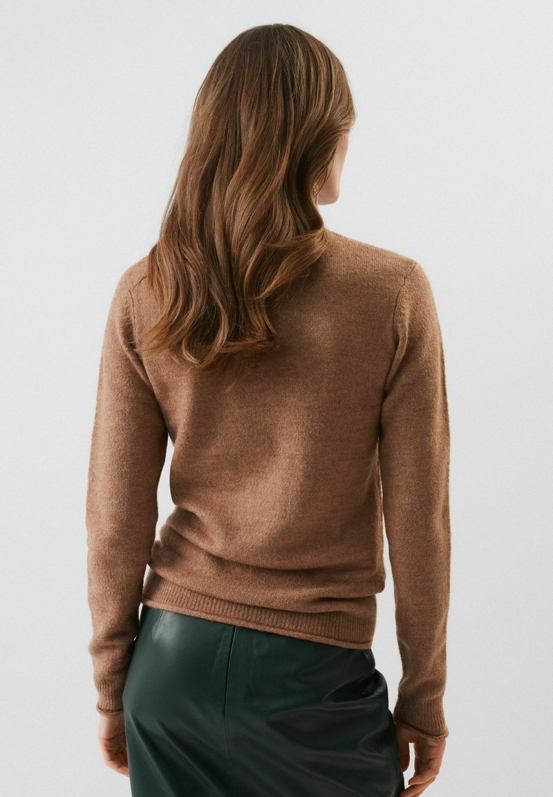 Femme avec de longs cheveux bruns portant un pull marron et une jupe en cuir vert foncé, se tenant face à un fond blanc uni.