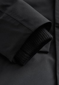 Sort jakke stof med en glat, mat tekstur; har en ribbet sort strikket manchet, der går fra ærmet for ekstra varme.