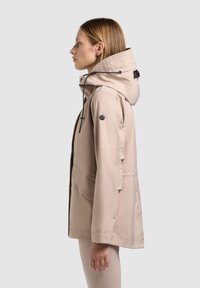 Naine küljelt, kannab beežikat kapuutsiga jopet, millel on reguleeritavad nöörid ja esitaskud, seisab ühevärvilise helge tausta ees.