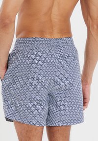 Costumi da bagno blu e bianchi con una fascia elasticizzata in vita, caratterizzati da un design geometrico e tasche laterali. Visuale posteriore mostrata.