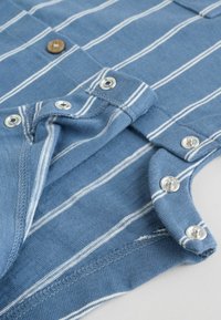 Tutina azzurra per bambini con righe bianche, dotata di bottoni a pressione lungo le gambe e un bottone in legno sulla parte superiore del petto.
