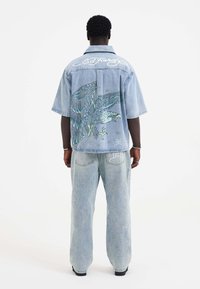 Denim kortærmet skjorte i lyseblå med ørn grafiktryk på ryggen, med broderet "Ed Hardy" på nakkestykkerne og afslappet pasform.