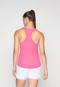 Mulher com penteado em rabo de cavalo trançado, vestindo um top de desporto sem mangas cor de rosa e calções atléticos brancos, de costas contra um fundo plain.