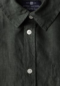 Mörkgrön linneskjorta med knappar, klassisk krage, vita knappar och en bröstficka, med en texturerad tyg yta.