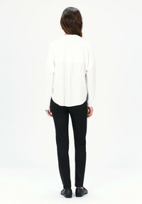 Weißes Langarmhemd mit abgerundetem Saum, kombiniert mit schwarzen enganliegenden Hosen und flachen schwarzen Schuhen, von hinten betrachtet.