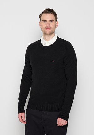 Uomo con capelli castani corti che indossa un maglione nero sopra una camicia bianca con colletto, in piedi davanti a uno sfondo grigio chiaro uniforme.