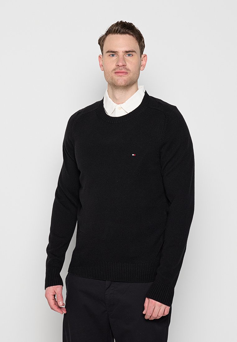Homme aux cheveux bruns courts portant un pull noir sur une chemise blanche à col, debout devant un fond gris clair uni.