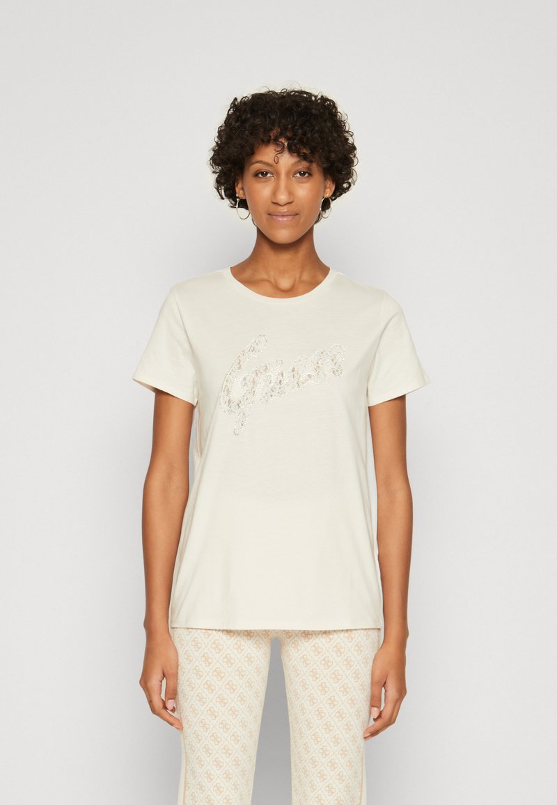 Camiseta de color beige claro hecha de una tela suave, con un diseño del logo "Guess" brillante. Manga corta y escote redondo.